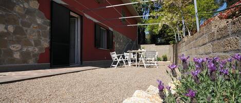 Un edificio con fachada de piedra y pared exterior roja. Cuenta con un patio con mesa y sillas, y un jardín con flores púrpuras.
