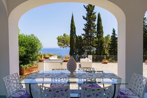 A glass-top table with white chairs and a decorative vase, set in an outdoor patio area with a view of the sea and tall trees. - Villa La Baika by MyVillaCollection (Capri)