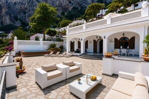 A sunlit outdoor seating area with a pool, white tiled flooring, and a view of a mountainous landscape. - Villa La Baika by MyVillaCollection (Capri)