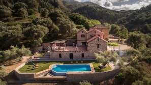 A stone building with a swimming pool, surrounded by trees and hills.