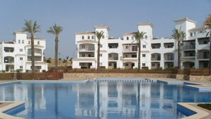 A swimming pool with a stone-tiled deck, surrounded by white multi-story buildings with balconies and palm trees. - 2 Bed Apartment On PGA Golf Course (Sucina)
