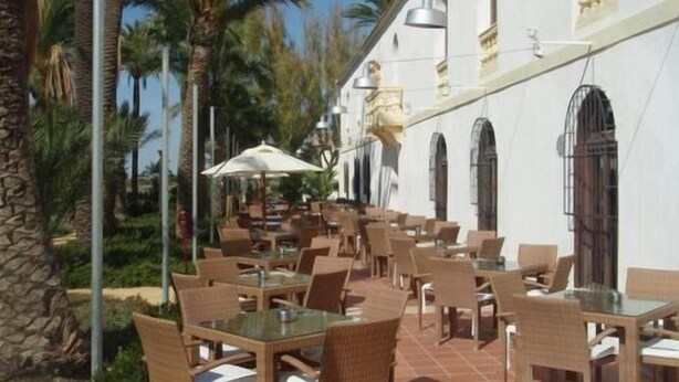 An outdoor cafe with wicker tables and chairs, a white building with arched windows, and palm trees in the background. - 2 Bed Apartment On PGA Golf Course (Sucina)