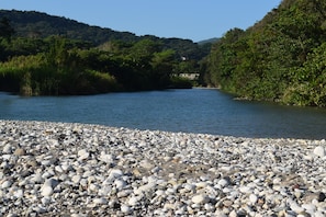 Orilla de río pedregosa con vistas a un bosque en la colina y un edificio distante.
