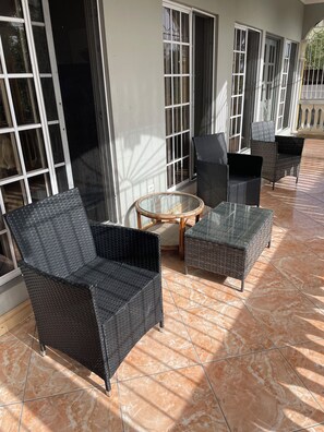 Un espace extérieur aménagé, abrité et doté de meubles en osier et d’une table en verre, situé à côté d’un bâtiment avec de grandes fenêtres.
