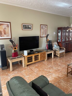 A living room with a flat-screen TV, wooden entertainment unit, green sofa, and a dining area with a table and chairs. - A superb 3-bedrooms Holiday Villa, mins away from Ocho Rios vibrant Town Centre (Tower Isle)