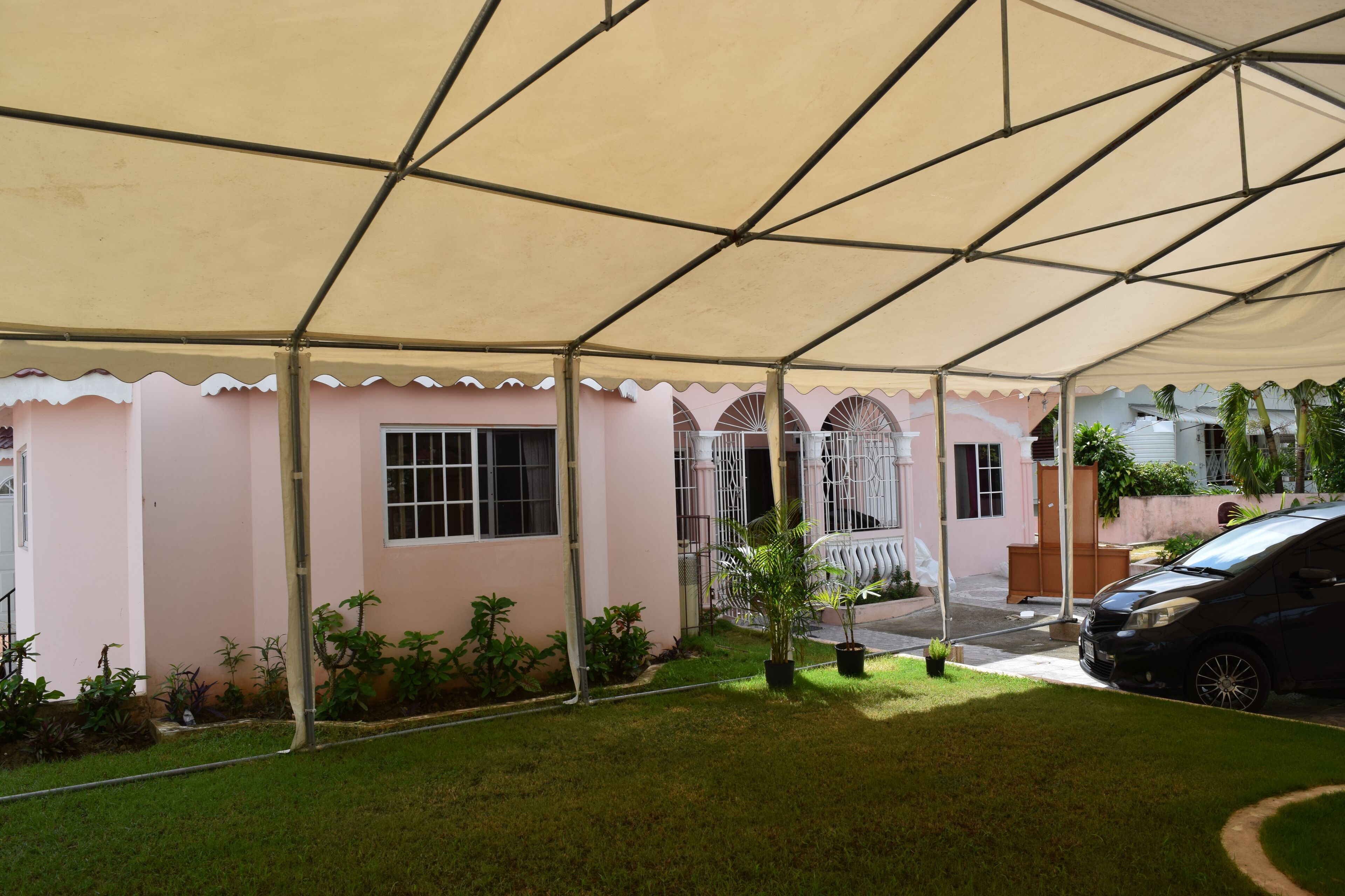 Un espace extérieur abrité, doté d’une structure en forme de tente, avec une voiture garée à proximité et un bâtiment à la façade rose en arrière-plan.