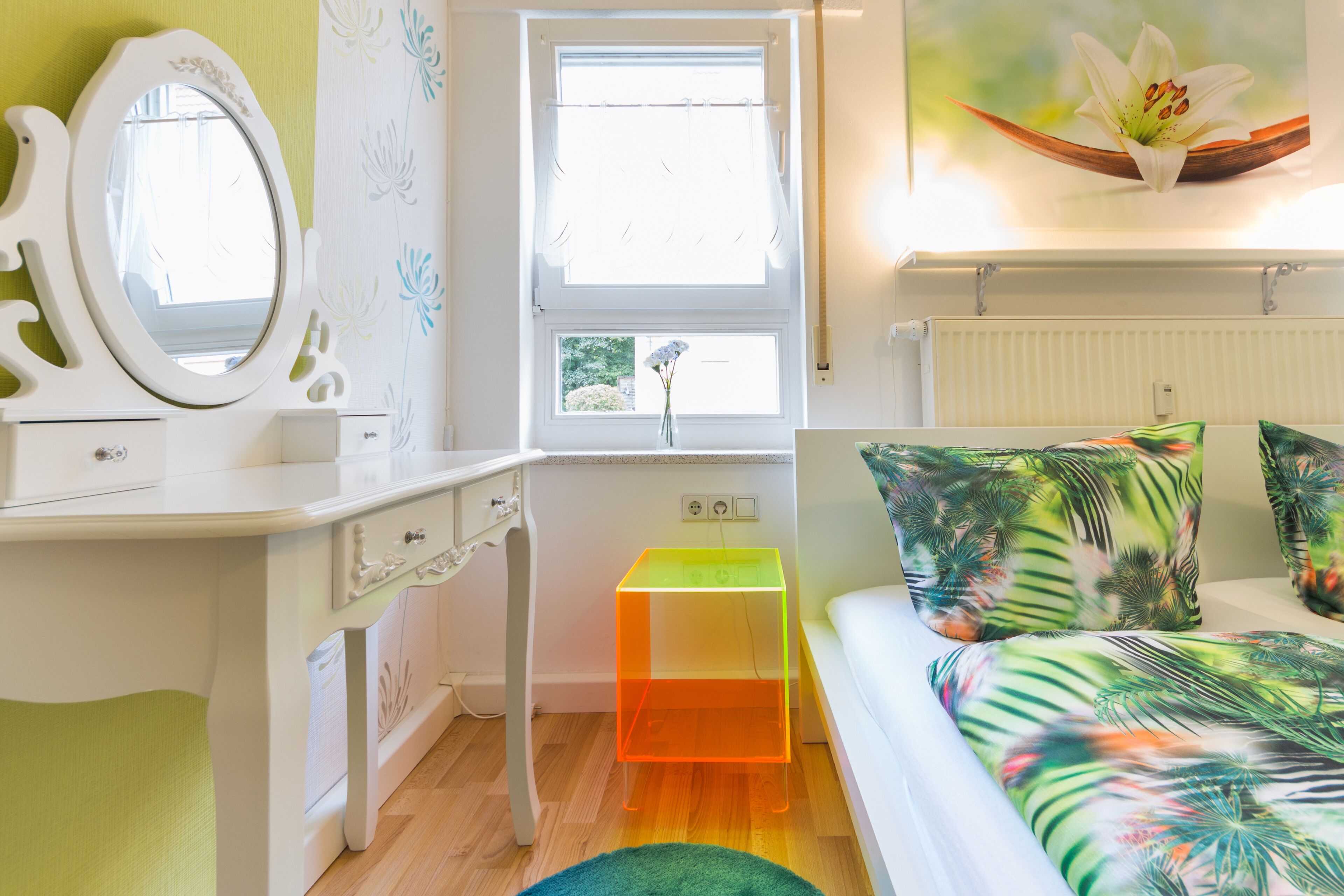 Ein Schlafzimmer mit Bett, Schminktisch, Spiegel, Fenster mit Blick und Heizkörper.