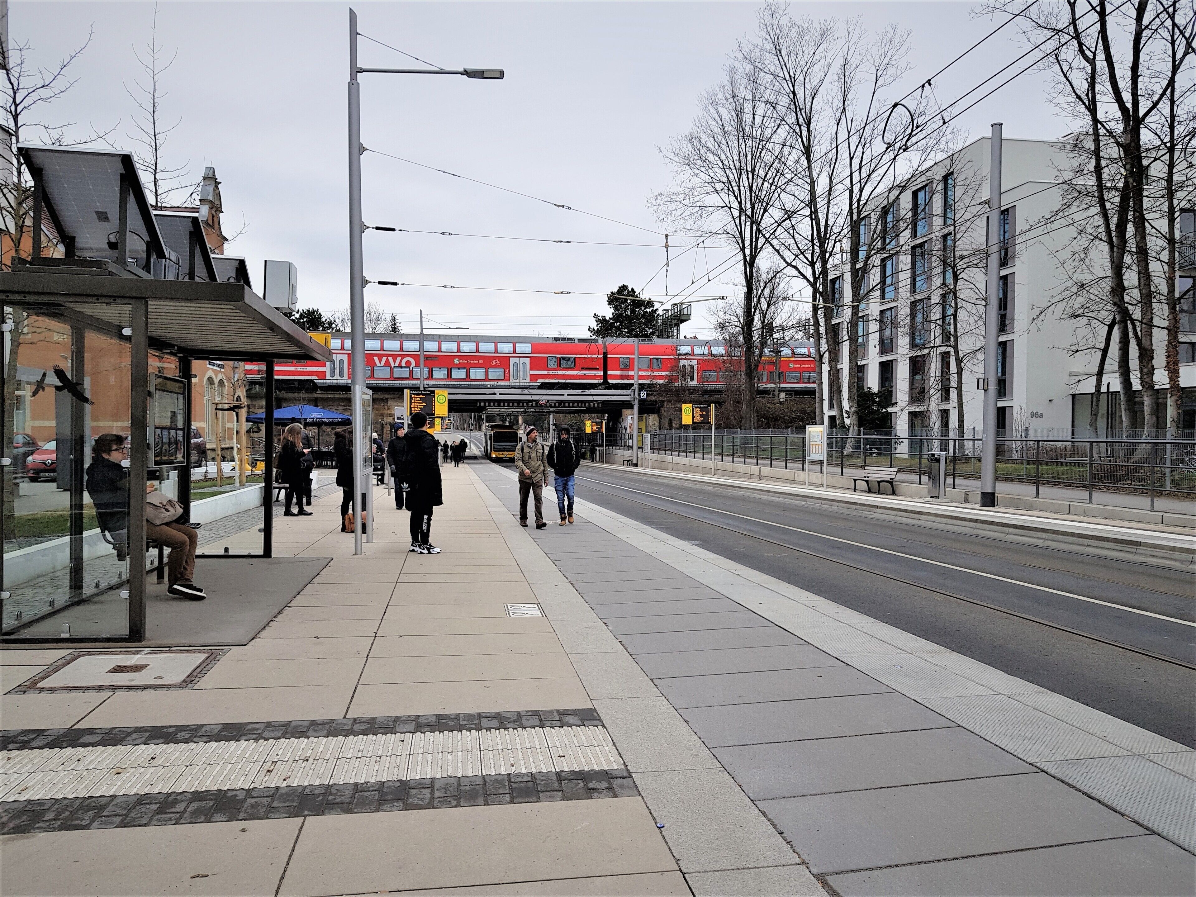 Ein Bahnsteig mit einem roten Zug im Hintergrund, einer Bushaltestelle und einem Fußgängerüberweg.