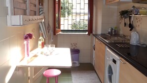 Cocina compacta con lavadora, una pequeña mesa de comedor con taburetes, ventana con vistas y estante con utensilios de cocina.