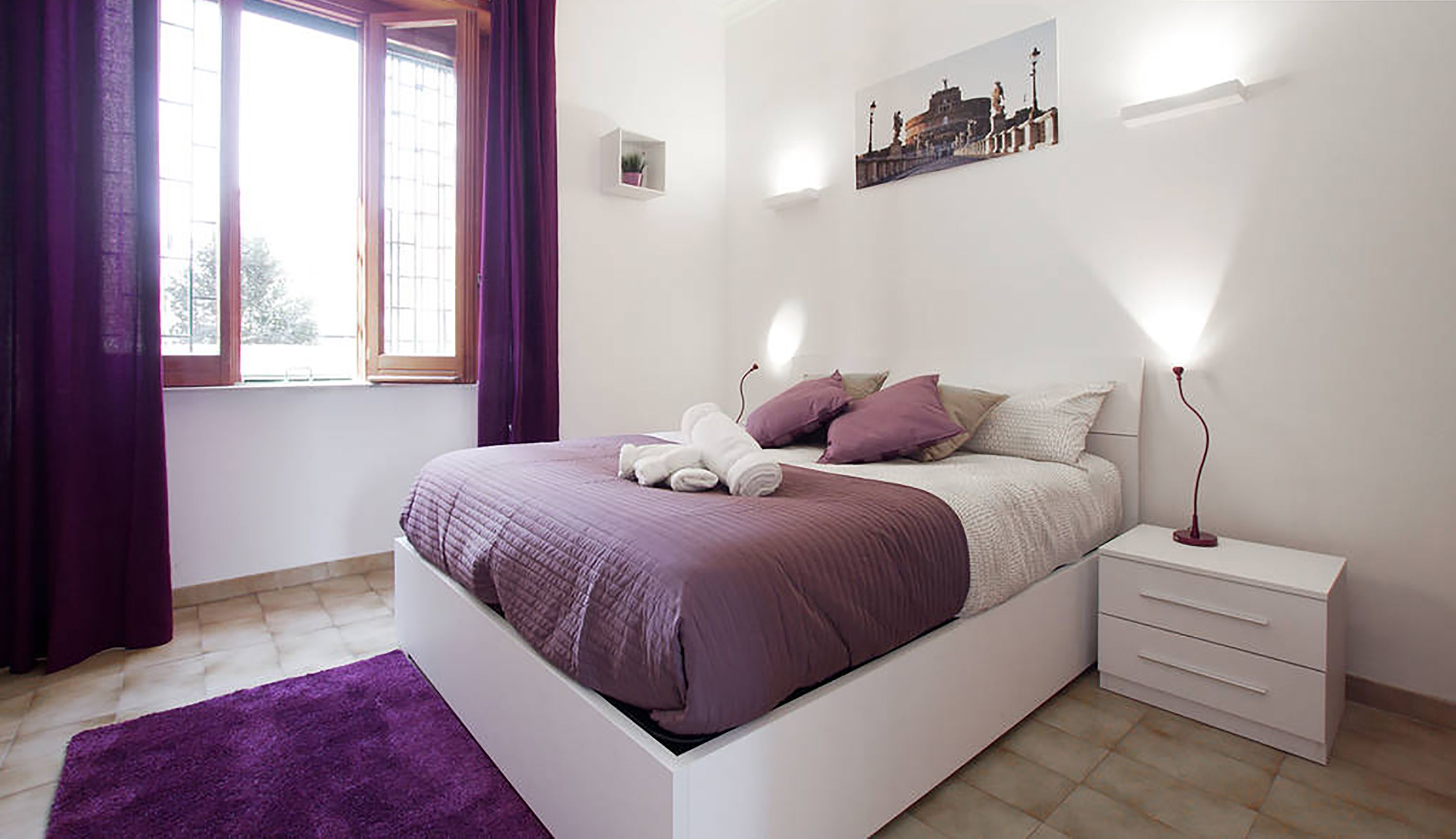 A bedroom with a bed, a nightstand, a lamp, a window with curtains, and a wall-mounted picture.