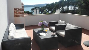 Un balcon agrémenté de meubles en osier, une table ornée de fleurs et sur laquelle se trouve une boisson, et une vue sur la mer et les arbres.