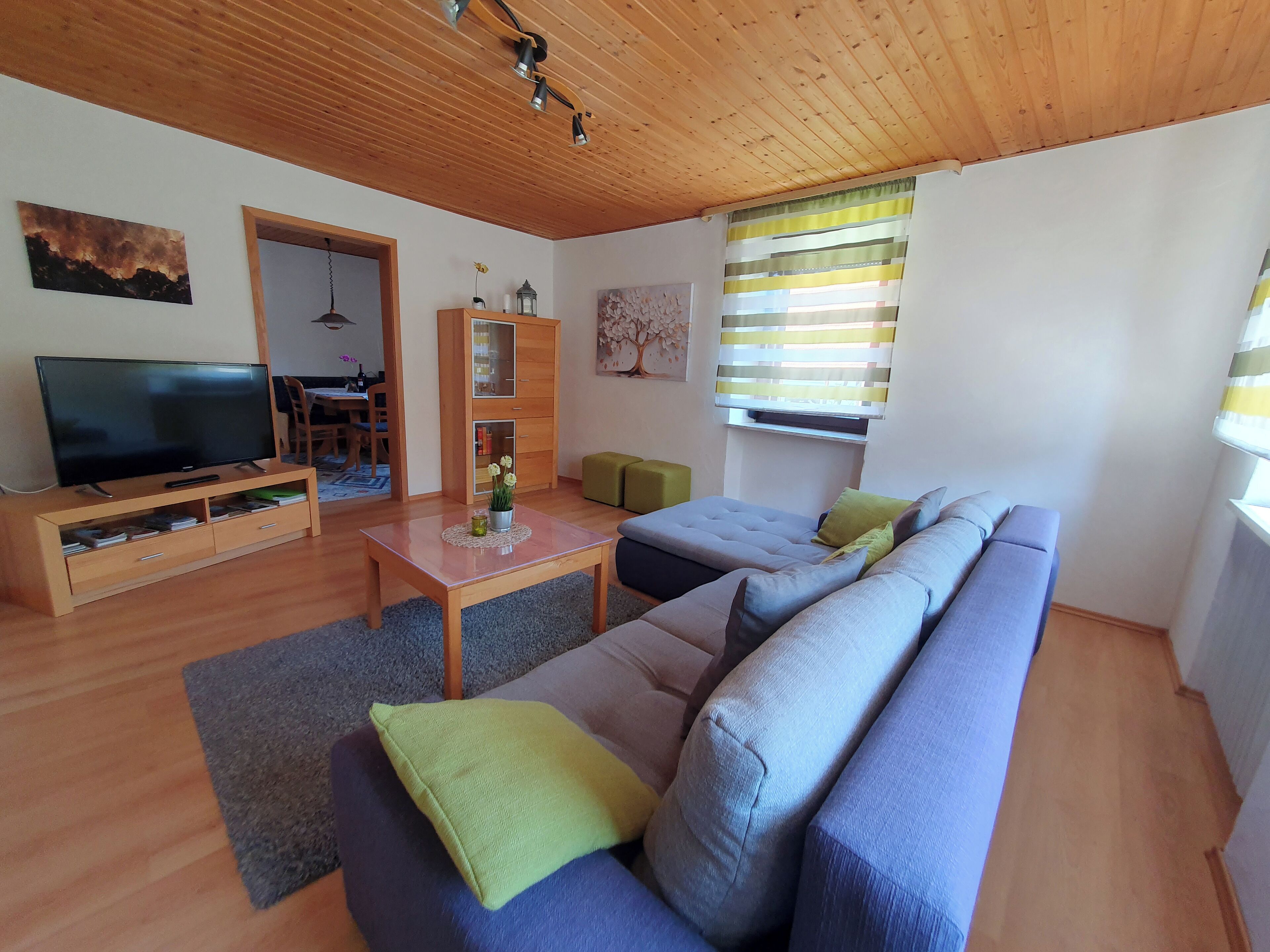 A modern living room with a wooden ceiling, a flat-screen TV, a wooden cabinet, a sofa, and a window with striped curtains.