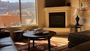 A living room with a fireplace, a sofa, a coffee table, and a view of a mountainous landscape through a large window. - 1A4 Shared Pool, Private Patio, Garage, Close to Arches (Moab)