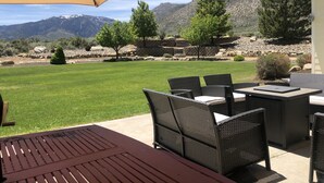 Un patio al aire libre con mobiliario de mimbre y un paraguas grande, con vistas a un césped cuidado y montañas.