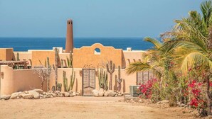 Paisaje desértico con un edificio que tiene una chimenea alta, cactus y palmeras.