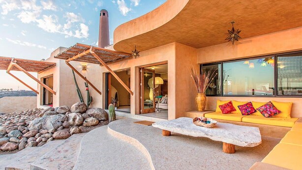 Un espacio exterior moderno con un edificio curvo, un banco de piedra, un sofá amarillo con cojines de colores y un ventanal grande.