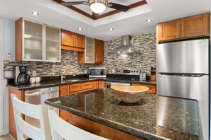 A modern kitchen with granite countertops, stainless steel appliances, wooden cabinets, and a central island with a bowl. - Experience paradise in our cozy 2 bedroom condo near the beach (Kihei)