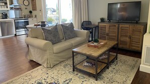Ein Wohnzimmer mit Sofa, Couchtisch, Fernseher, Holzschränken und einer Küche im Hintergrund.