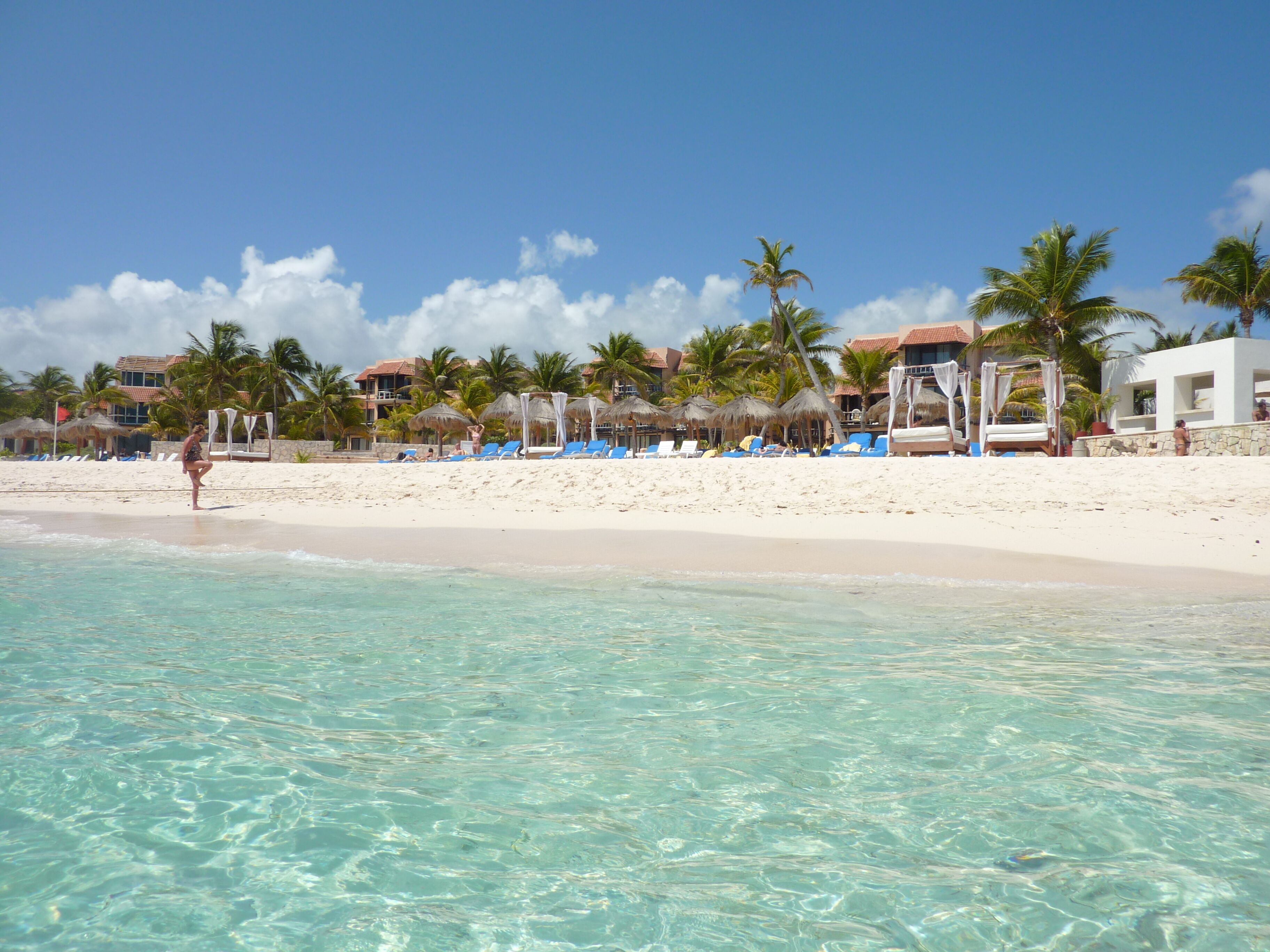 A beach with clear turquoise water, white sand, and palm trees. There are blue lounge chairs and umbrellas along the shore.
