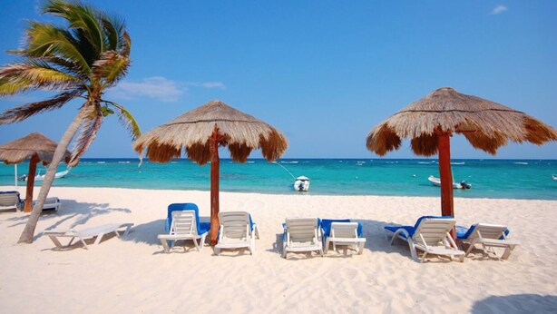 Une plage de sable blanc, sous un ciel bleu, avec une eau turquoise. On y trouve des parasols en chaume et des chaises longues.