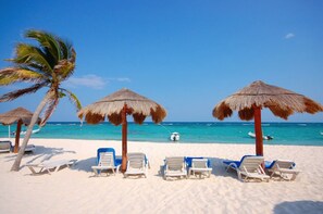 Une plage de sable blanc, sous un ciel bleu, avec une eau turquoise. On y trouve des parasols en chaume et des chaises longues.