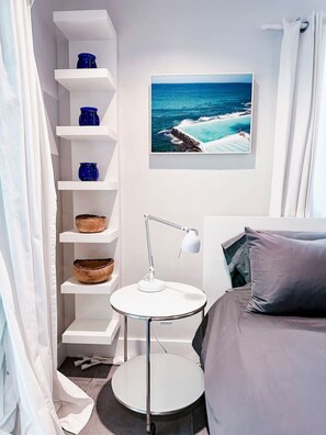 A bedroom with a bed, a round table with a lamp, a shelf with decorative items, and a framed picture of a beach scene.