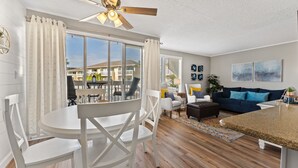 A modern living room with a dining area, large windows, a ceiling fan, and a view of neighboring buildings. - One of SPC's most booked units! Poolfront! Brand NEW renovation!  (Destin)