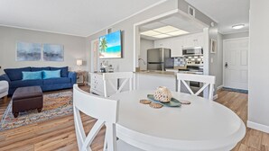 A modern living room with a blue sofa, a round dining table with white chairs, and a kitchen in the background. - One of SPC's most booked units! Poolfront! Brand NEW renovation!  (Destin)