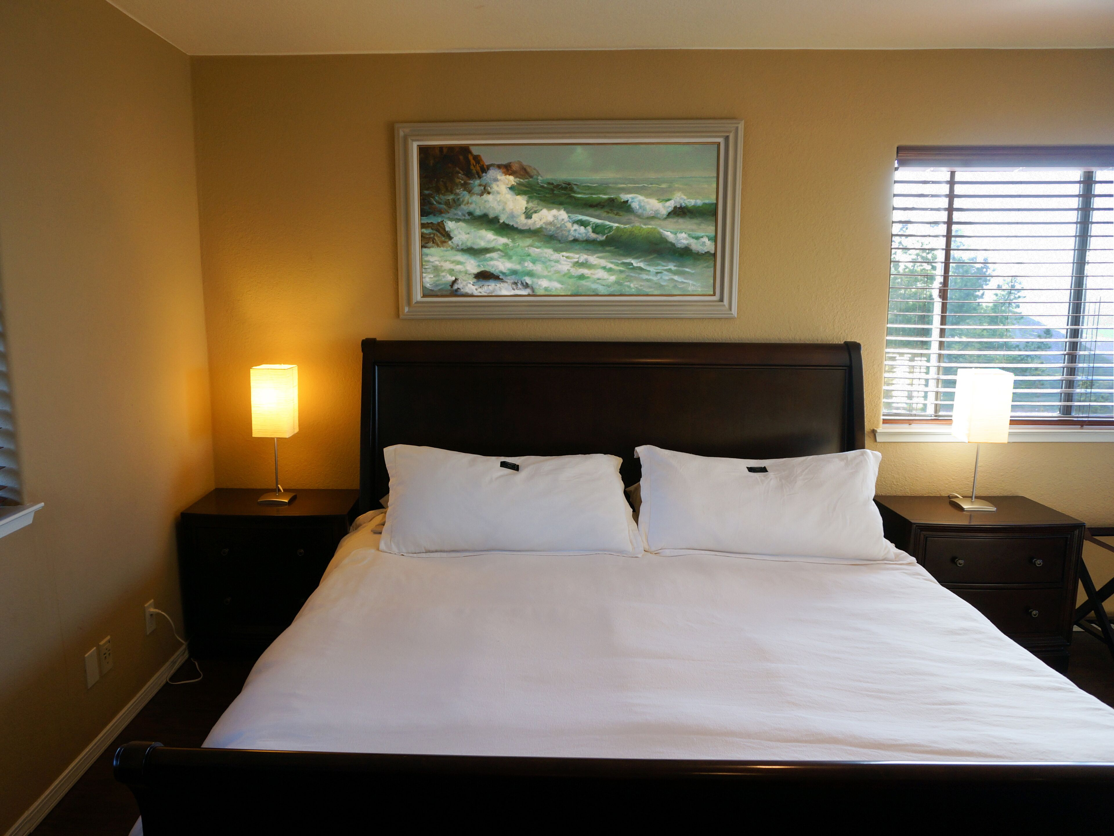 Habitación de hotel con cama, dos mesitas de noche, un cuadro de olas y una ventana con persianas.