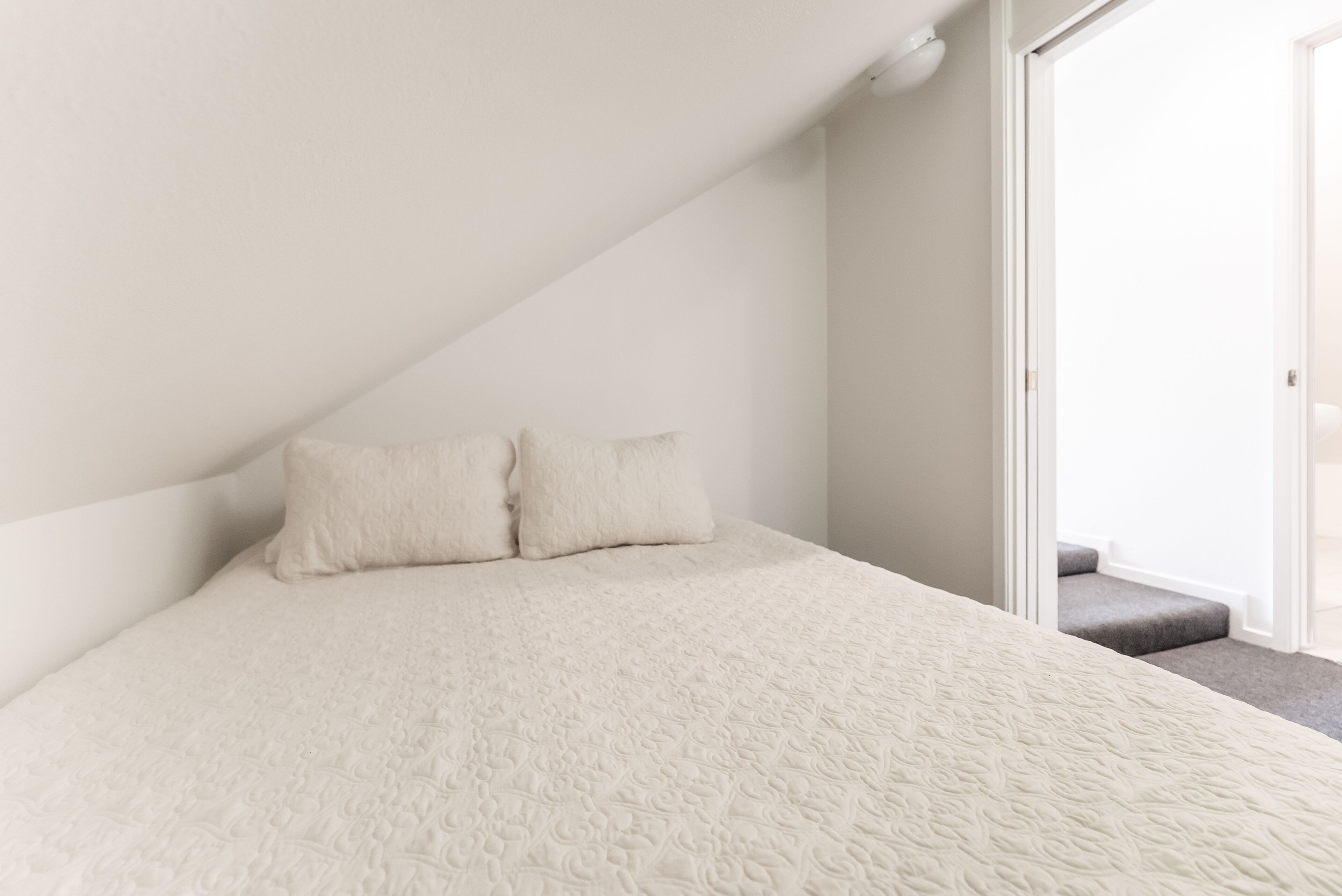 Une chambre avec un plafond en pente, un lit recouvert d’une courtepointe blanche à texture, deux oreillers et une porte donnant sur un balcon.