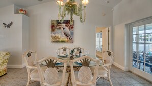 Un comedor con una mesa de vidrio, sillas de mimbre, una araña y un cuadro de un pájaro, además de una escultura de pájaro en la pared.