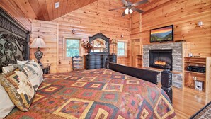 Ein Schlafzimmer in einer Blockhütte mit einem großen Bett, einem Kamin, einem Deckenventilator und einem Fernseher, der über dem Kamin angebracht ist.