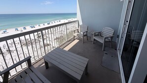 A balcony with a view of the beach, a table, two chairs, and a bench.
