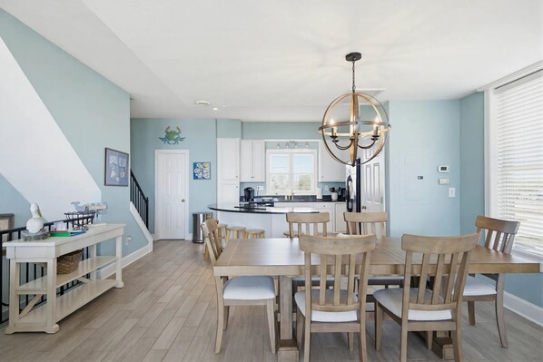 Un coin repas avec une table et des chaises en bois, un lustre et, en arrière-plan, une vue sur la cuisine.