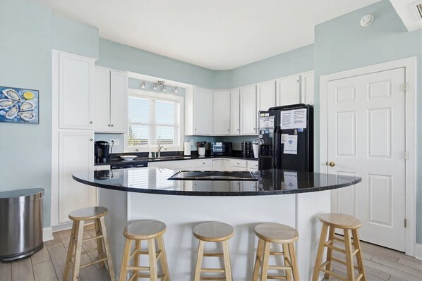 A kitchen with white cabinets, a black refrigerator, a large island with stools, and a window with blinds. - 🌞Oceanfront 🏖️Steps to sand 🐶Dog Friendly⚡️EV charger (North Topsail Beach)