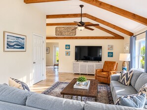 A spacious living room with a ceiling fan, a flat-screen TV, and a large window with curtains.