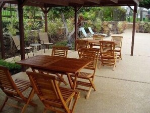 An outdoor patio area with wooden tables and chairs, surrounded by greenery and a covered structure.