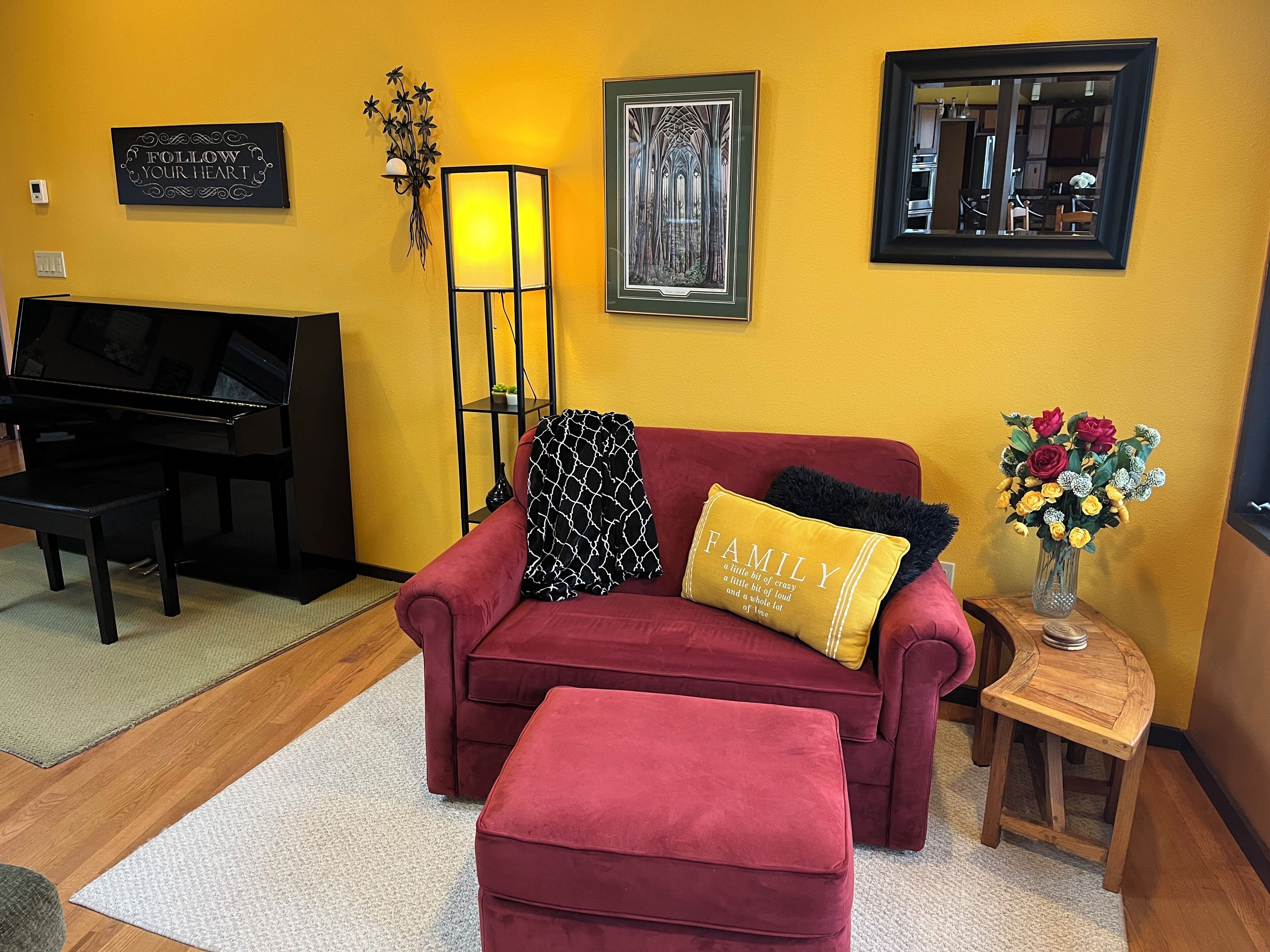 A living room with a red sofa, a matching ottoman, a black piano, a framed artwork, a wall-mounted lamp, and a vase of flowers.