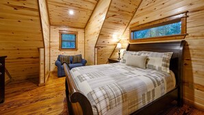 Un accogliente sottotetto adibito a camera da letto, con soffitto spiovente in legno, un letto grande con copriletto a quadri, un comodino, una lampada, una finestra vista alberi e un divano blu.