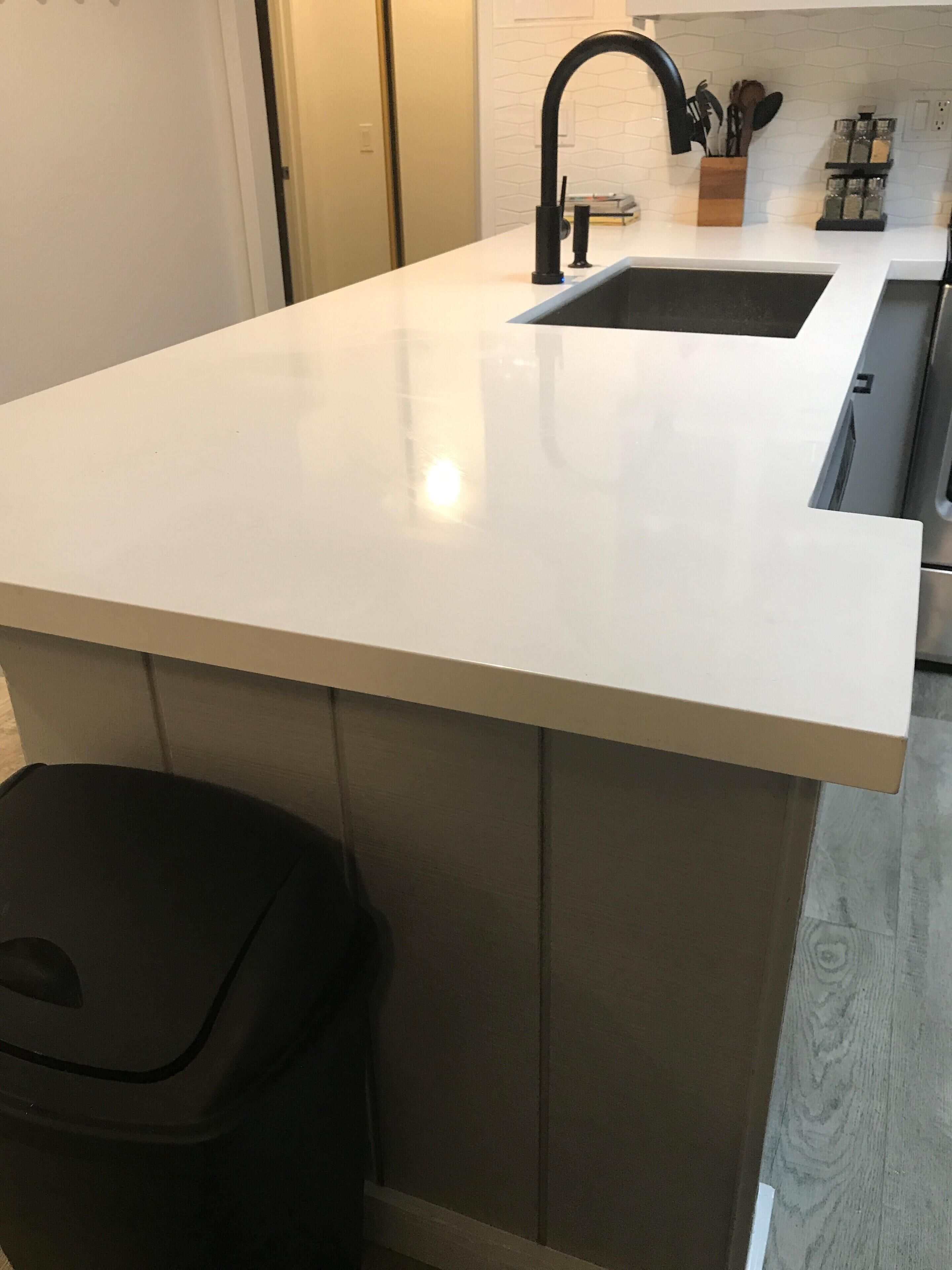 Una cocina con encimera blanca, un cubo de basura negro y un grifo negro.