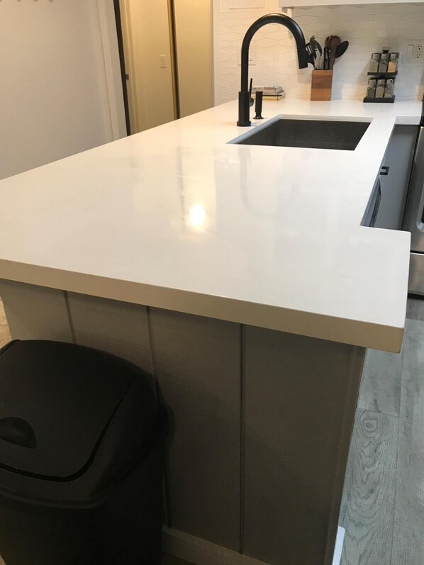 A kitchen with a white countertop, a black trash bin, and a black faucet.