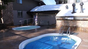 Two outdoor swimming pools with steps leading into the water, a stone wall, and a building with a snow-covered roof in the background.
