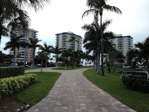 A paved pathway leading to a building complex with multiple floors, surrounded by palm trees and greenery. - 7 Miles of Paradise!  Completely Renovated Condo. Cleaning Included.   (Fort Myers Beach)