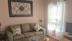 Ein Wohnzimmer mit einer Couch, zwei gemusterten Kissen, einem Glastisch, einer Wanddekoration, einer TV-Konsole und einem Fenster mit transparenten Vorhängen.