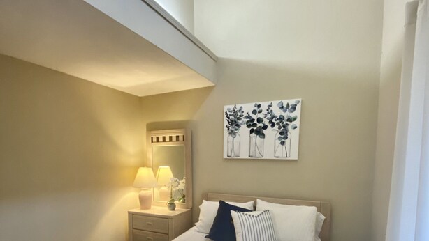 Ein ordentlich bezogenes Bett mit weißer Daunendecke und blauen Kissen, ein Nachttisch mit Lampe sowie ein an der Wand befestigtes Kunstwerk mit Vasen und Blumen.