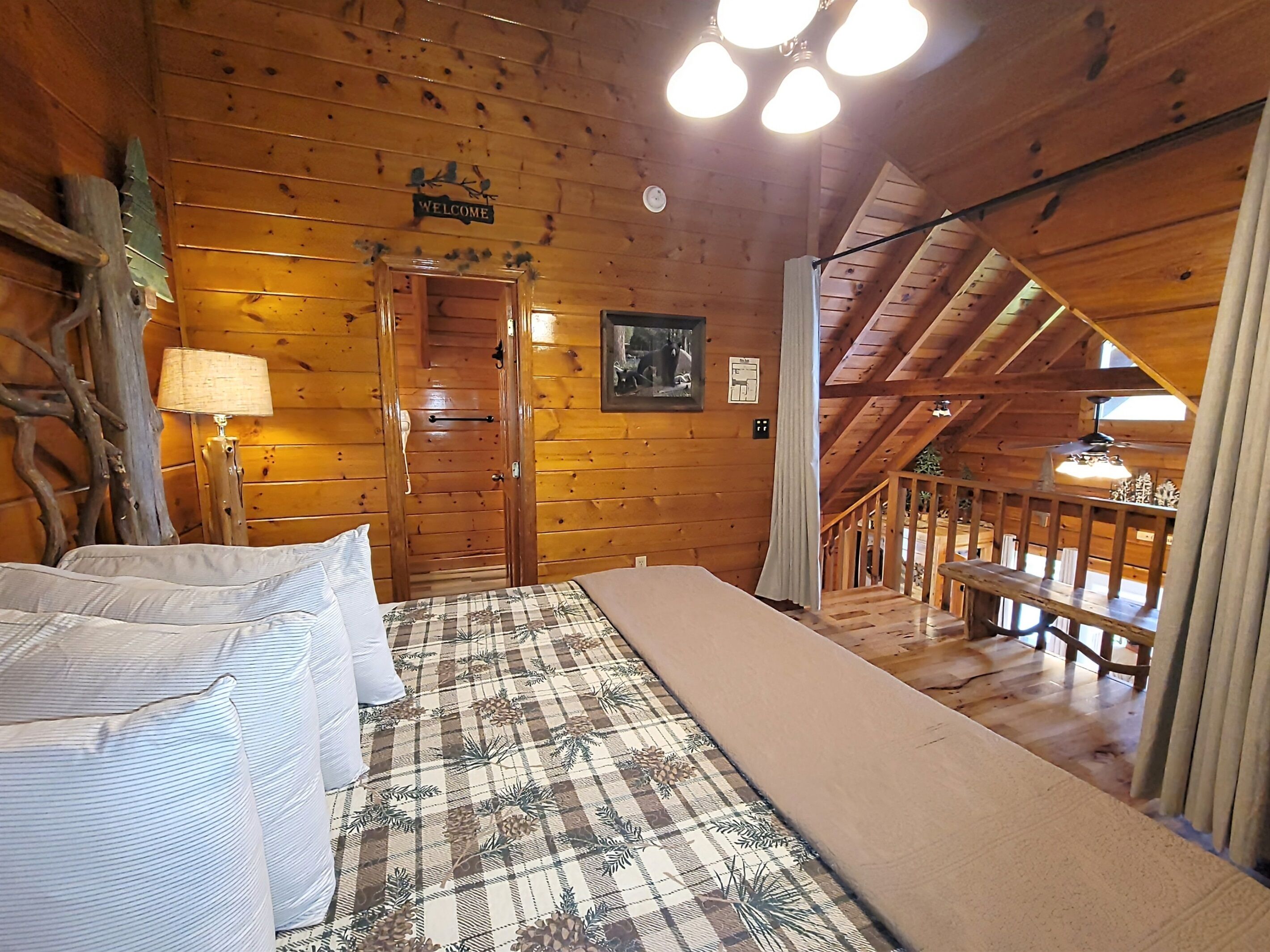 A cozy wooden cabin room with a bed, a welcome sign, a lamp, and a staircase leading to an upper level.