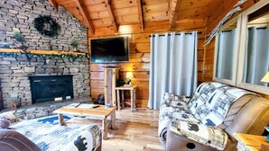 Ein gemütliches Blockhaus-Interieur mit Kamin aus Stein, einem an der Wand befestigten Fernseher und einer komfortablen Sitzecke mit Sofa und Sesseln.