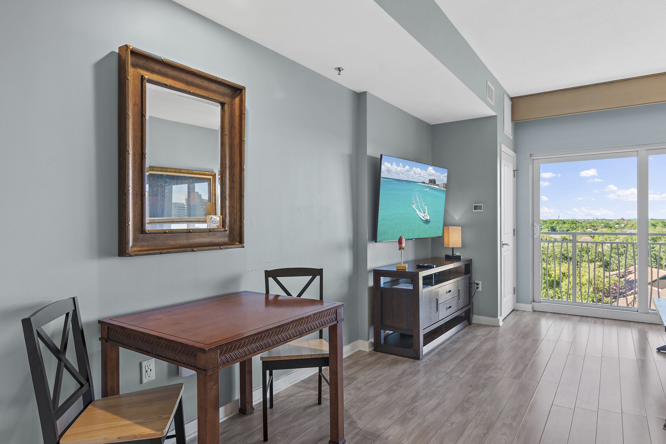 A dining area with a wooden table and chairs, a TV on a stand, and a balcony with a view.