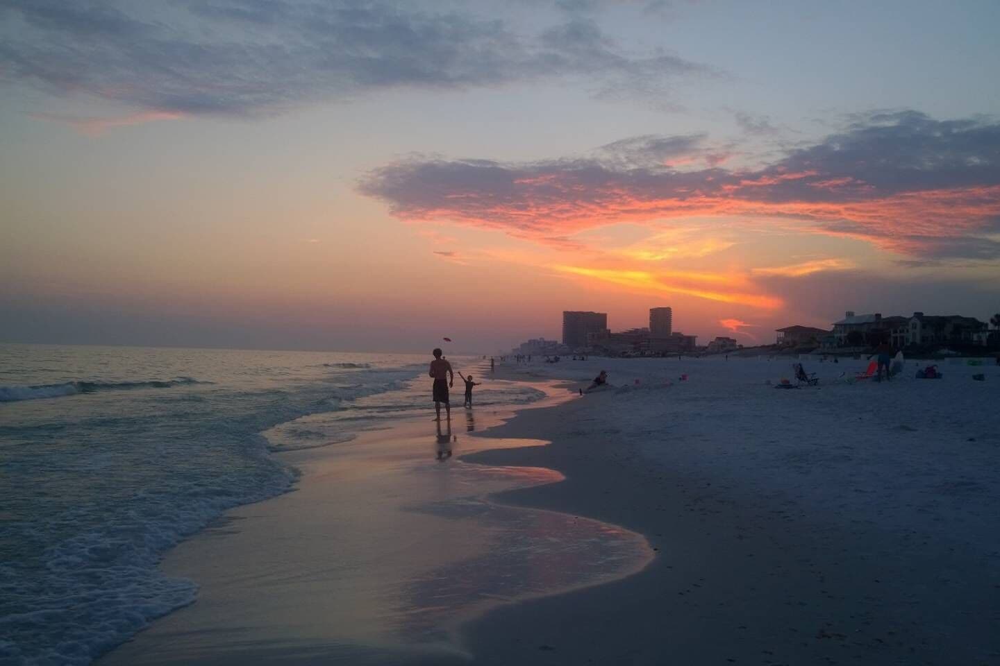 Eine Strandansicht mit Sonnenuntergang, Wellen und Gebäuden im Hintergrund.