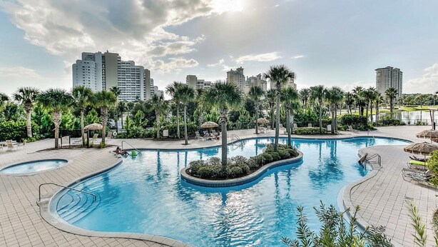 A large outdoor swimming pool surrounded by palm trees and lounge chairs, with high-rise buildings in the background.
